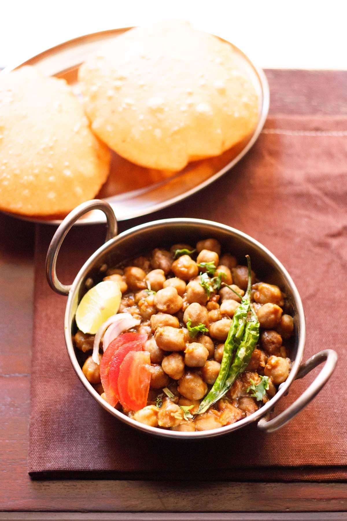 chana masala in a small serving wok topped with sliced tomatoes, onions, lemon and green chillies served with poori.