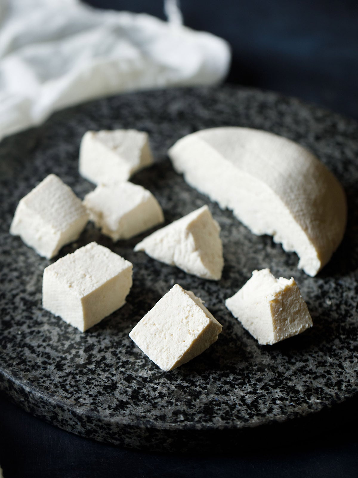 paneer block halved and cut in cubes on a grey-black granite round board – a guide on how to make paneer.