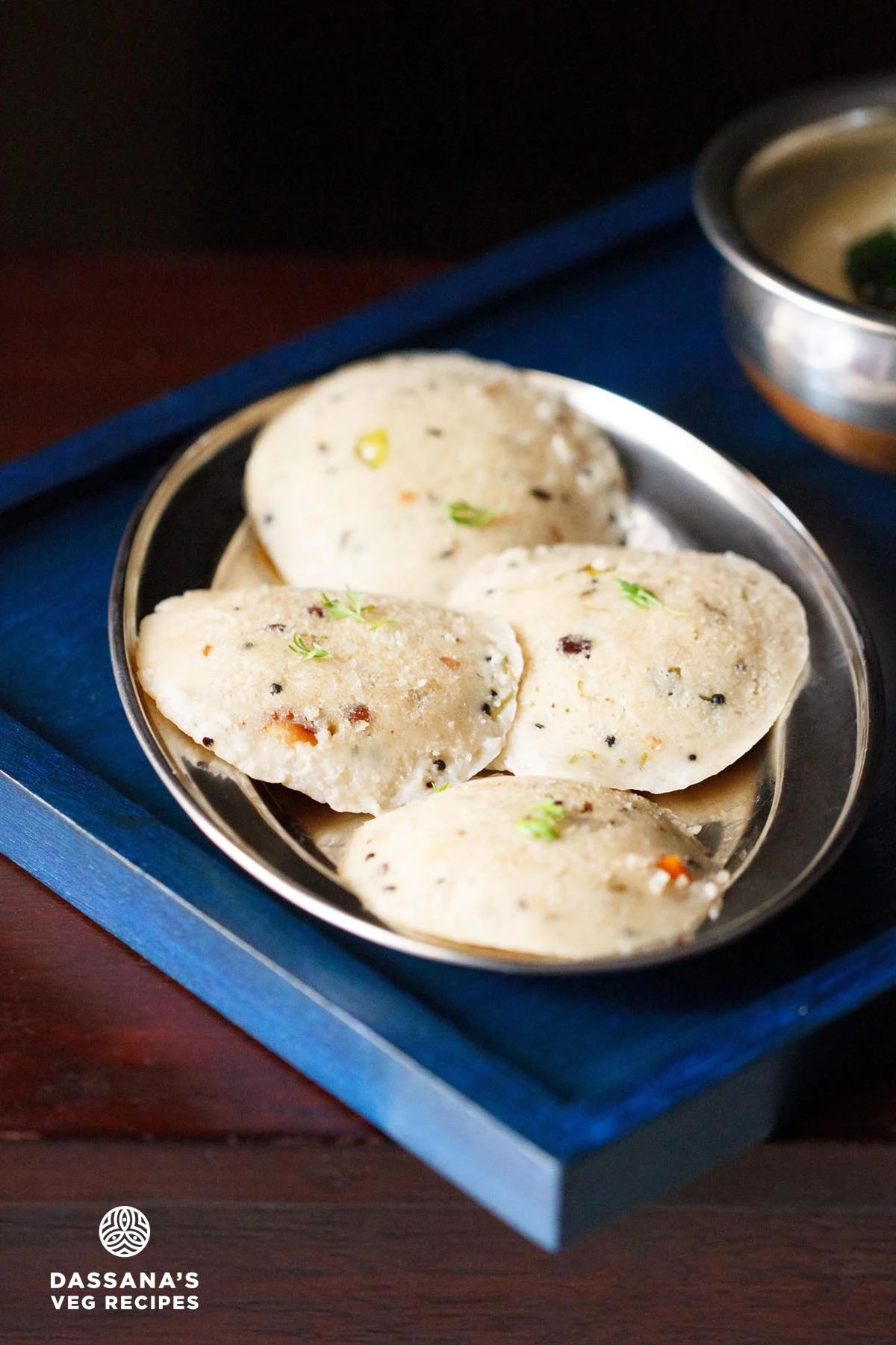 Four round oats idli arranged on silver plate on a dark blue wooden tray with coconut chutney in a silver bowl.