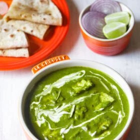 palak paneer topped with cream, garnished with ginger julienne and served in an orange bowl on a white board with roti kept on a dark orange plate on top left and a small orange bowl with round sliced onions and lemon wedges on top