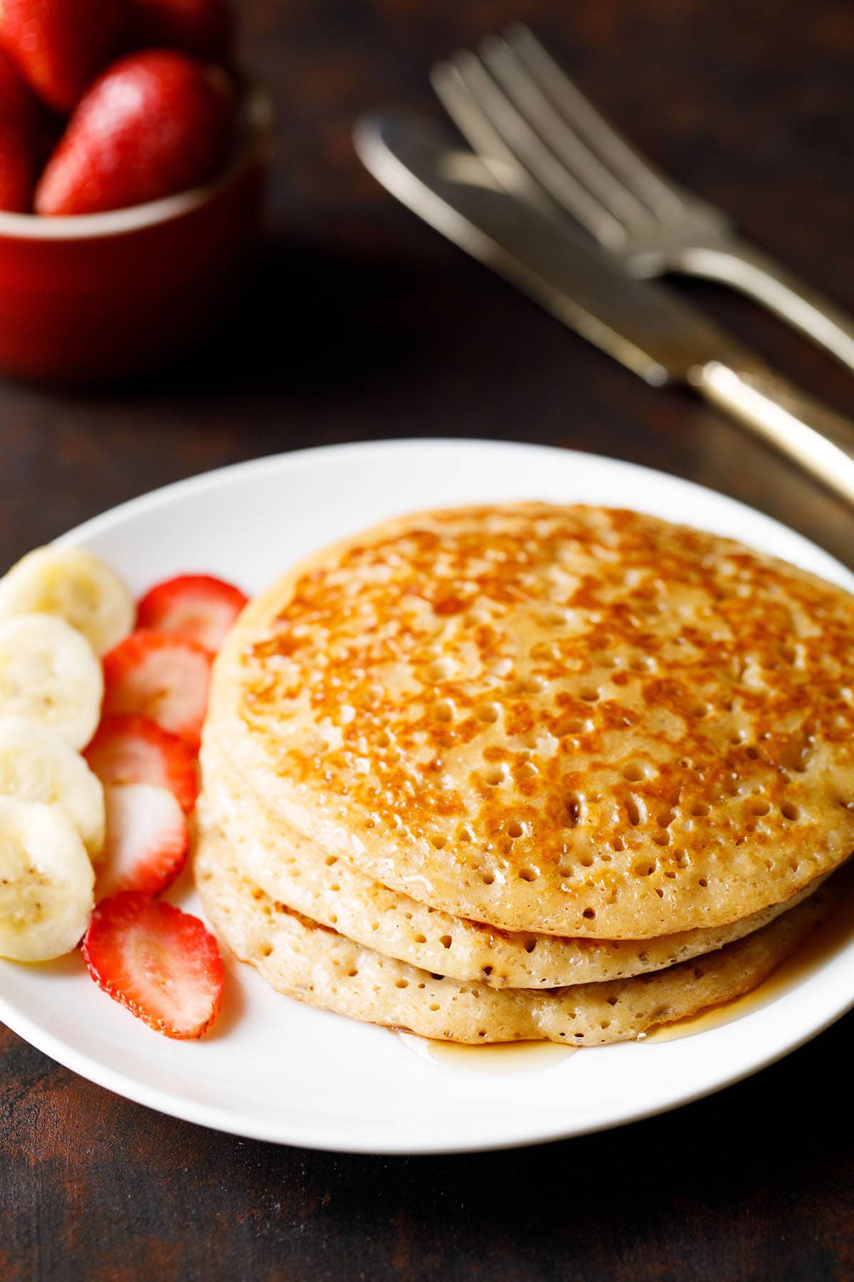 stack of eggless pancakes with maple syrup drizzled and sliced bananas and strawberries on white plate.