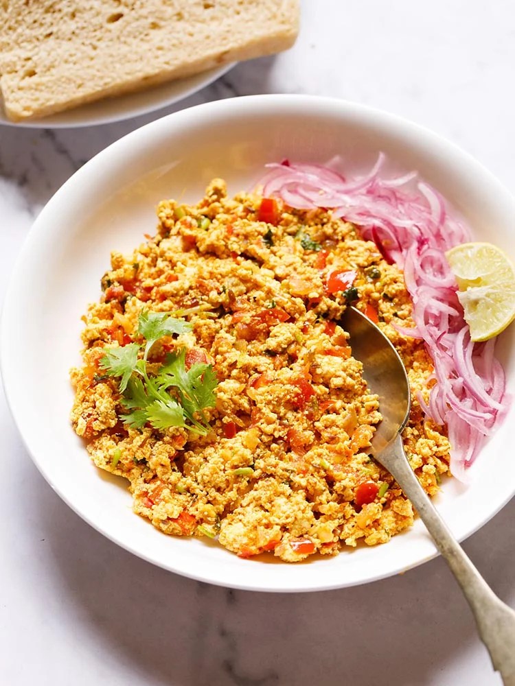 paneer bhurji garnished with a coriander leaf and served in a white plate with onion strips and lemon wedges kept on right side and a spoon inside it.