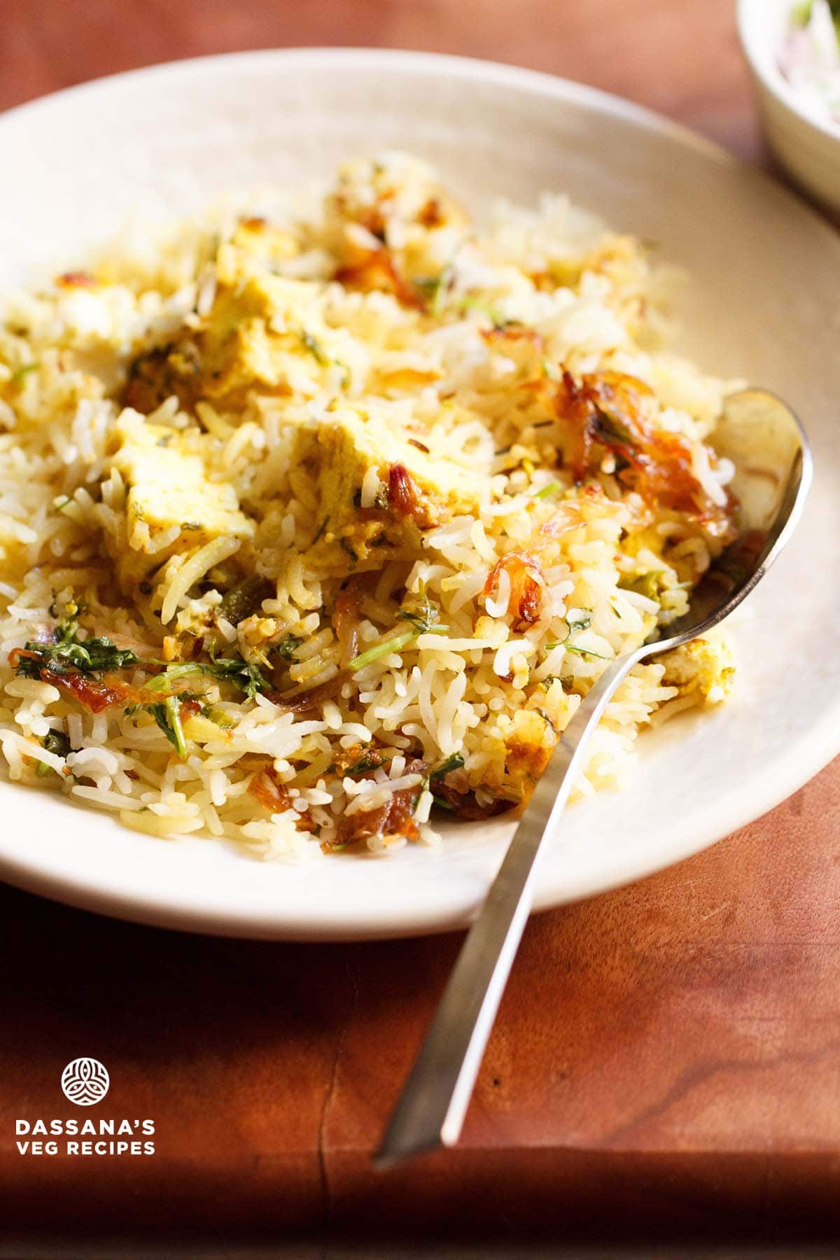 paneer biryani served in a cream colored shallow plate with a steel spoon by the side on a brown wooden board.