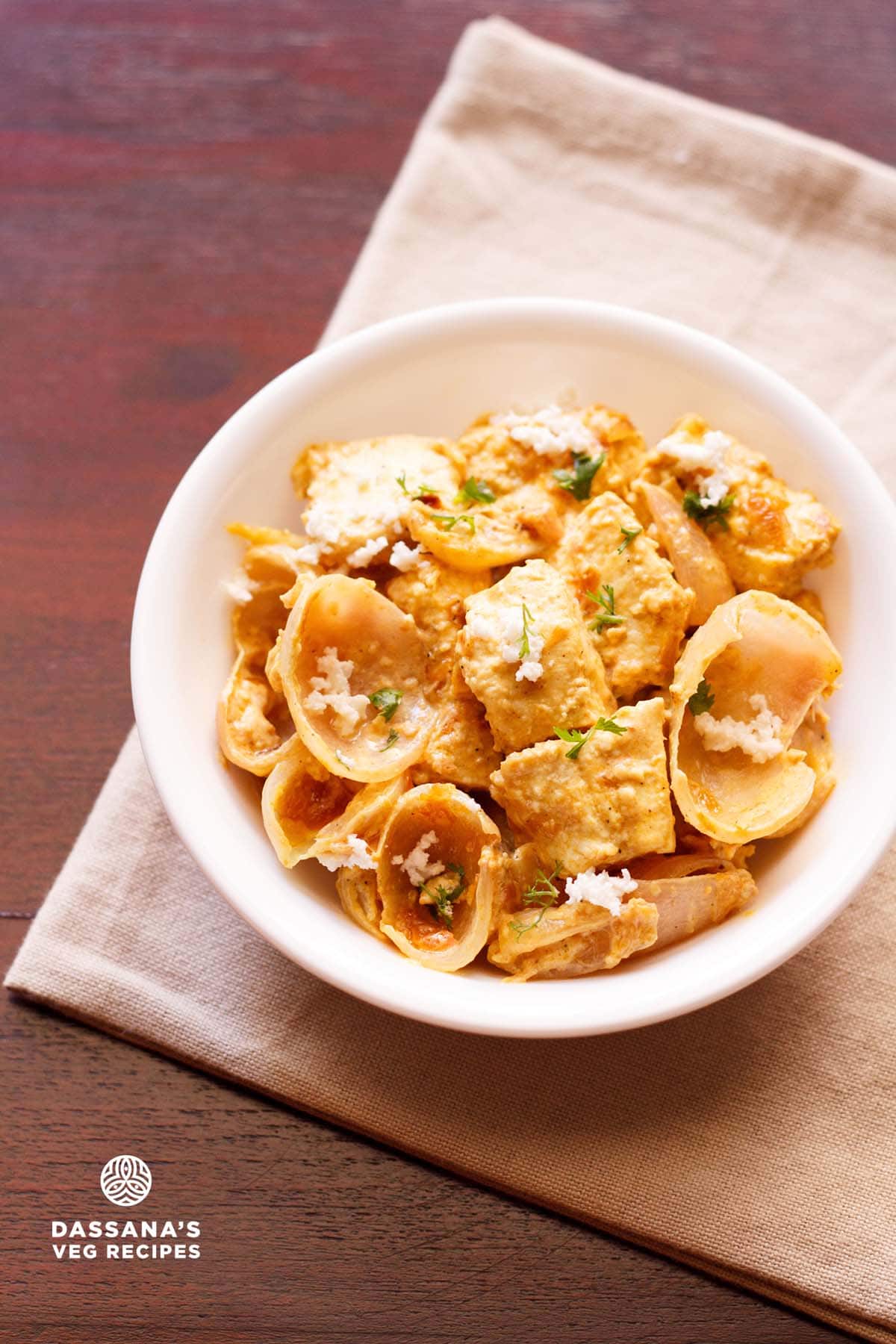 paneer do pyaza garnished with grated paneer and coriander leaves in a white bowl on a light beige napkin on a dark brown board.
